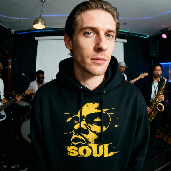 Man i Funky Soul hoodie svart framför liveband med gitarr och saxofon — musikinspirerad streetwear från Acceso.